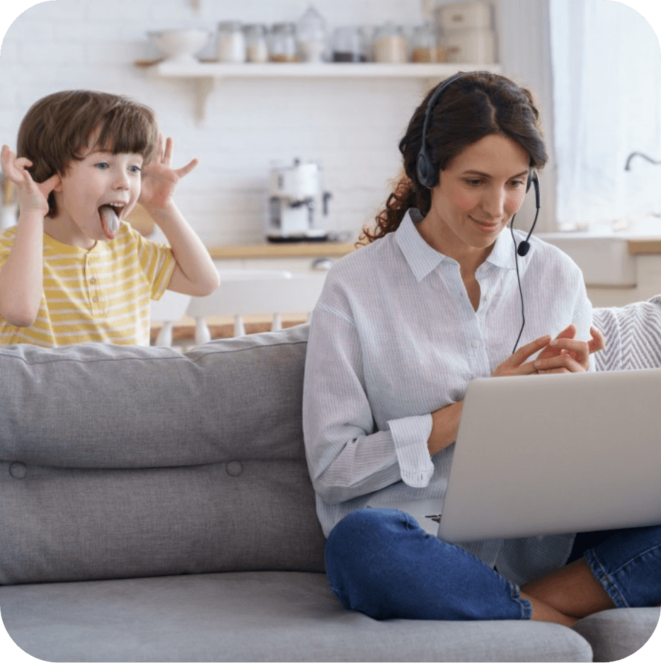 Employee Working at Home on Computer with Headset while Young Boy Makes Silly Face