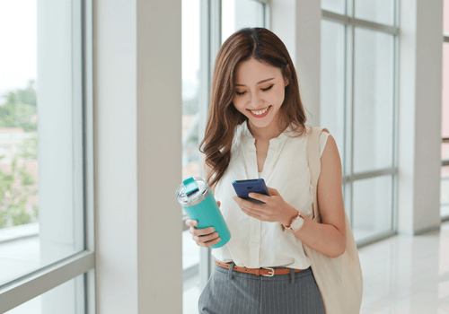 A woman holding a teal travel mug and blue smart phone in hallway off an office building