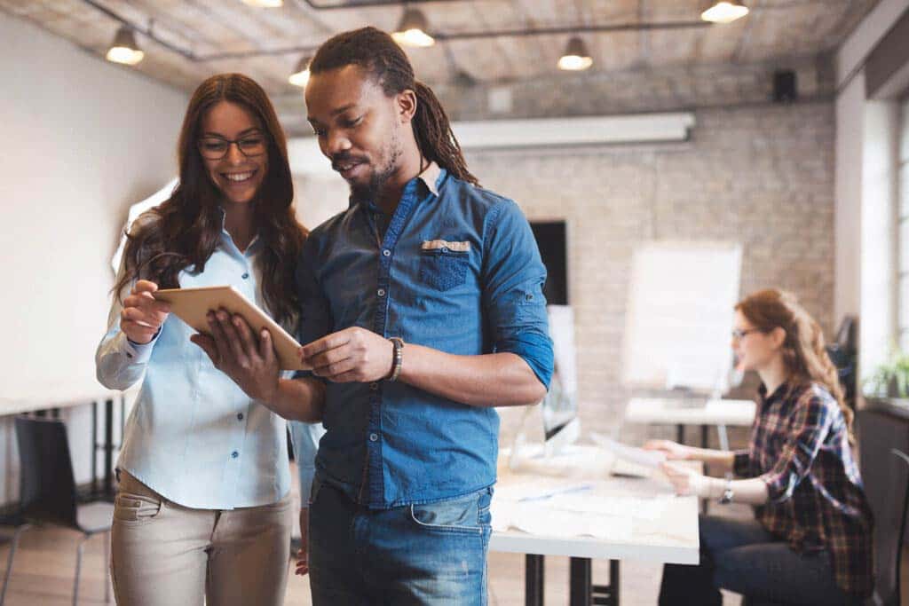 Man and Woman Standing Together in Office Looking at Notebook with Co-Worker in Background